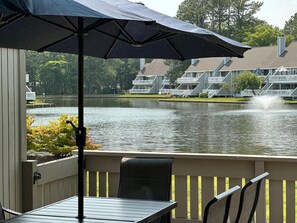 Vista al lago con una mesa y sillas en el patio, un parasol grande y un edificio con balcones al fondo.