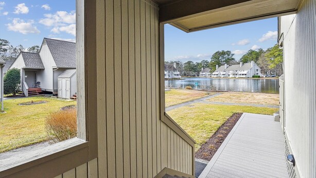 A view from a porch with a wooden railing, overlooking a grassy yard and a body of water with buildings in the background. - Lakefront Sea Colony Villa! Amenity Rich with Private Beach and Shuttle! (Bethany Beach)