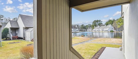 Une vue depuis une véranda dotée d’une balustrade en bois, donnant sur une cour verdoyante et un plan d’eau, avec des bâtiments en arrière-plan.