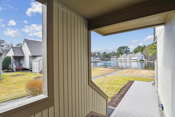 A view from a porch with a wooden railing, overlooking a grassy yard and a body of water with buildings in the background. - Lakefront Sea Colony Villa! Amenity Rich with Private Beach and Shuttle! (Bethany Beach)