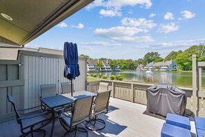 A covered outdoor area with a table and chairs, a covered grill, and a view of a lake with houses in the background.
