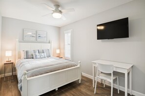 Un dormitorio con cama, mesitas de noche, escritorio, silla, ventilador de techo y televisor.