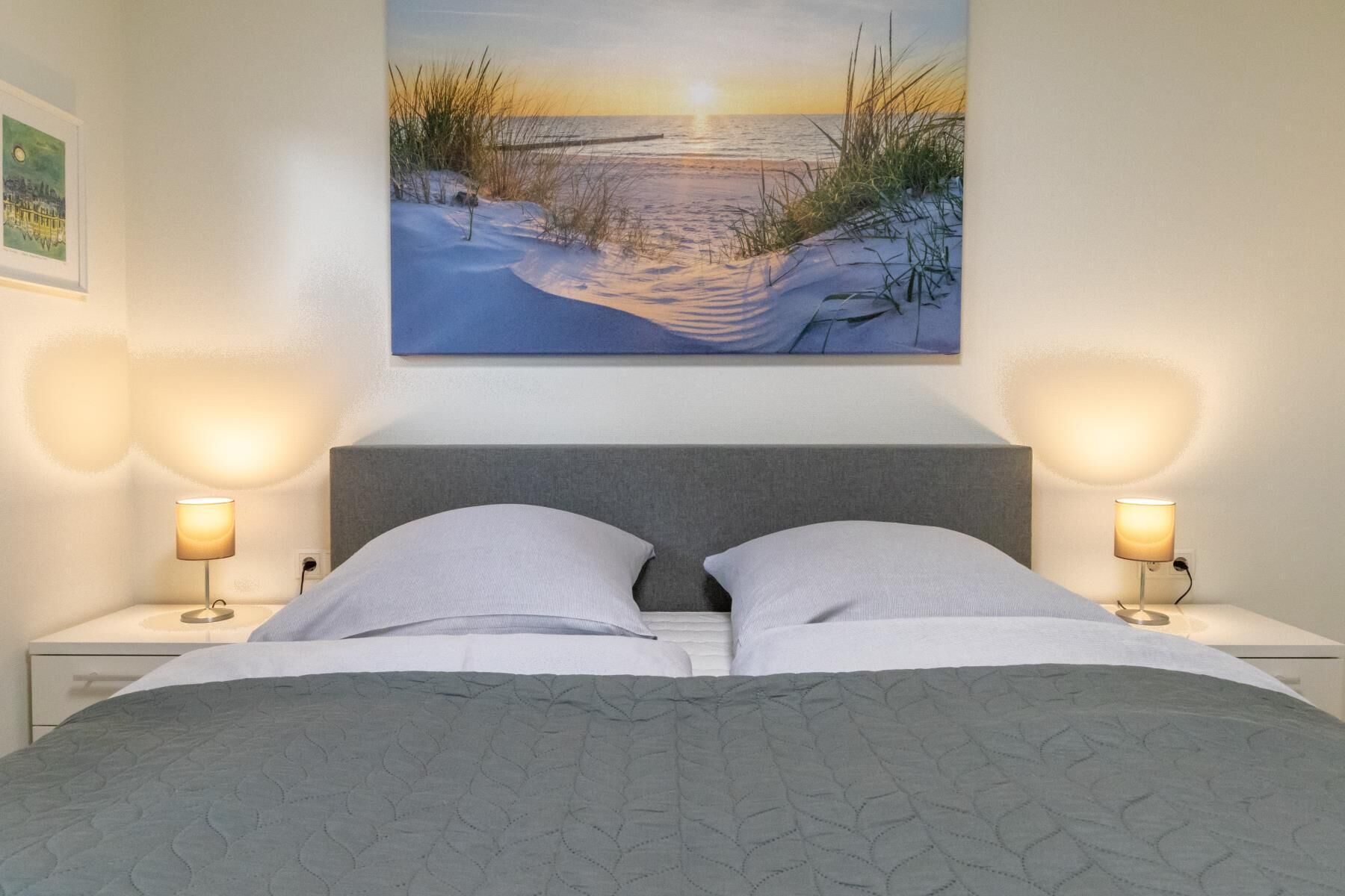 Ein ordentlich bezogenes Bett mit einer grauen Tagesdecke, zwei Nachttischlampen und einem Strandbild über dem Bett.