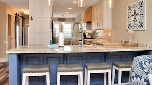 A kitchen with a marble island, bar stools, pendant lighting, and a decorative wall piece. - The Blue Pearl (Port St. Joe)