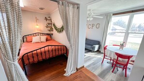 A bedroom with a bed, a desk, and a chair. There is a window with curtains and a view of the outside. - Oak Street Station in Downtown Mahomet (Mahomet)