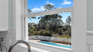 Vista desde una ventana con piscina y cerca blanca, que da a un jardín ajardinado con árboles y cielo despejado.
