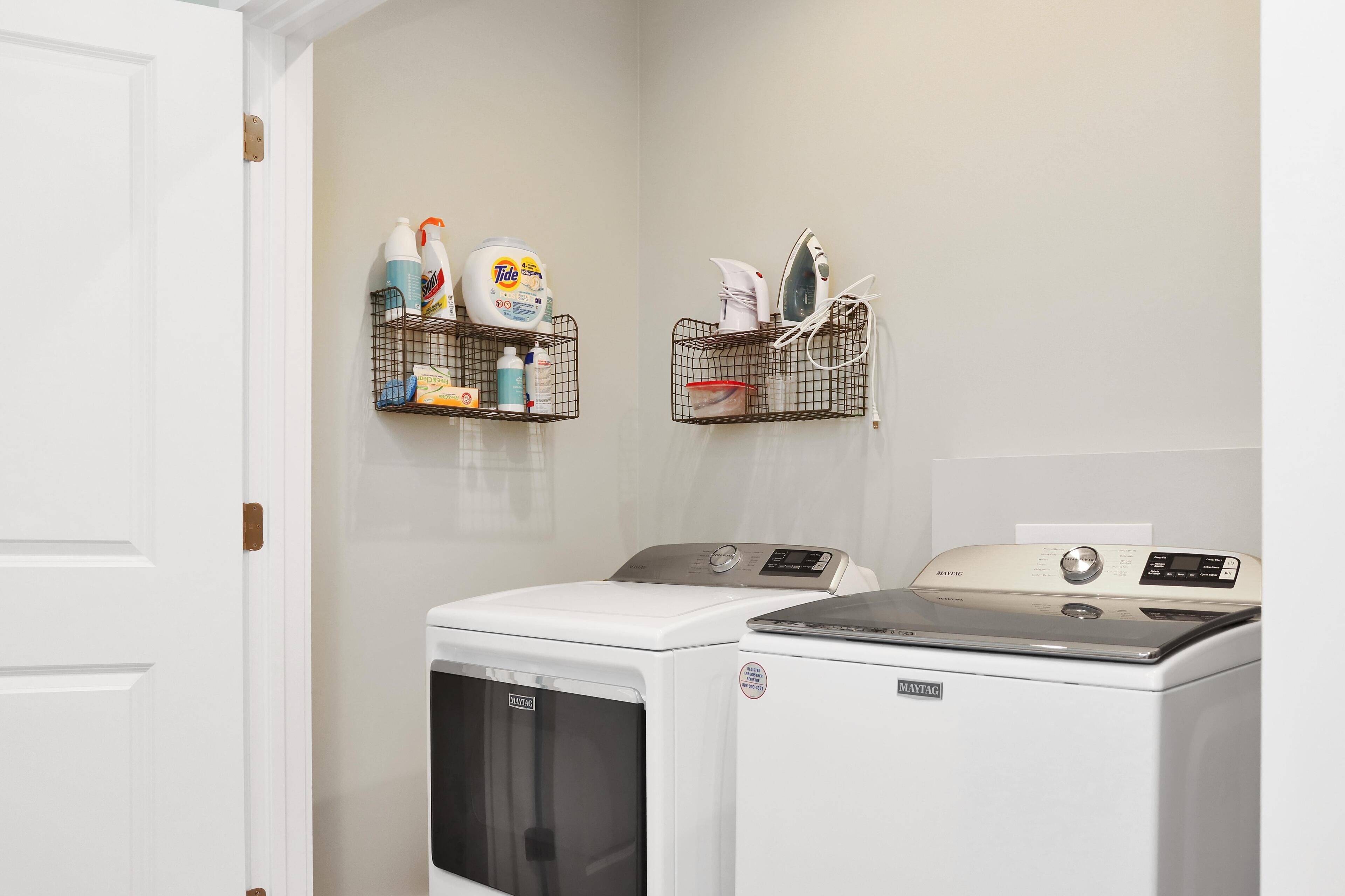 A laundry room with two washing machines, a shelf with cleaning supplies, and a wall-mounted ironing board.