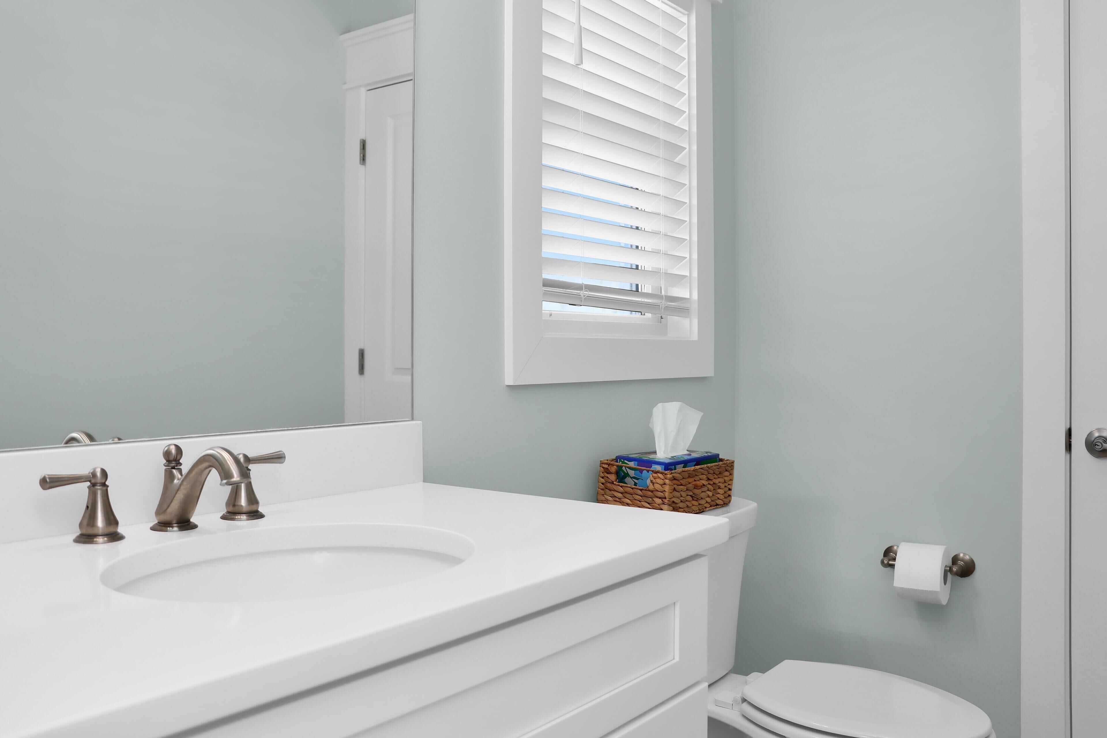 A bathroom with a white sink, two faucets, a mirror, a toilet, and a window with blinds.