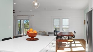 Una cocina moderna con encimera blanca, un cuenco de madera con frutas, una zona de comedor con sillas y un refrigerador.