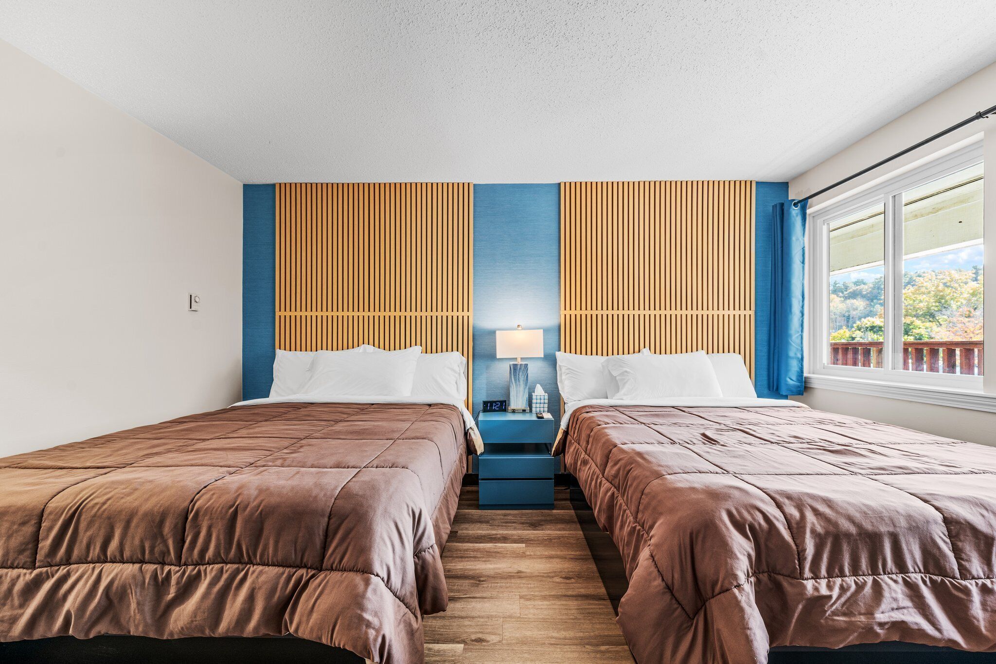 Ein Hotelzimmer mit zwei Betten, einem Nachttisch mit Lampe und einem Fenster mit Blick auf Bäume.