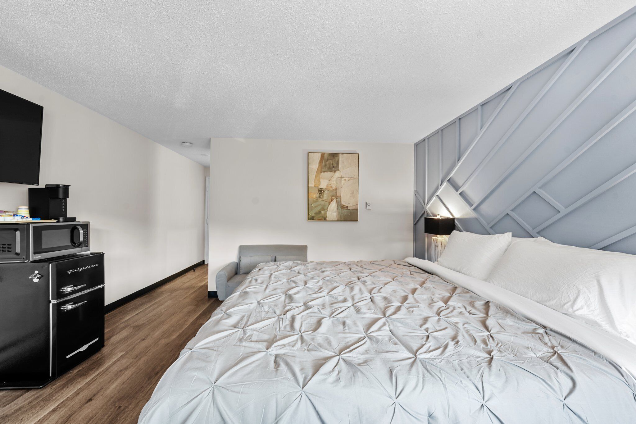 A modern bedroom with a large bed, a flat-screen TV, a coffee maker, and a painting on the wall.