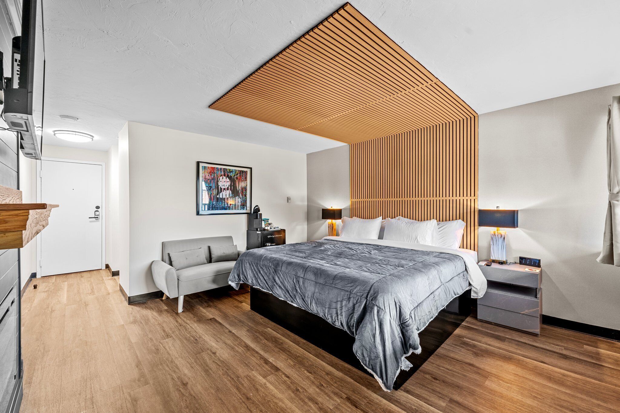 A modern bedroom with a large bed, a bedside table, a lamp, a gray sofa, and a wooden slatted ceiling.