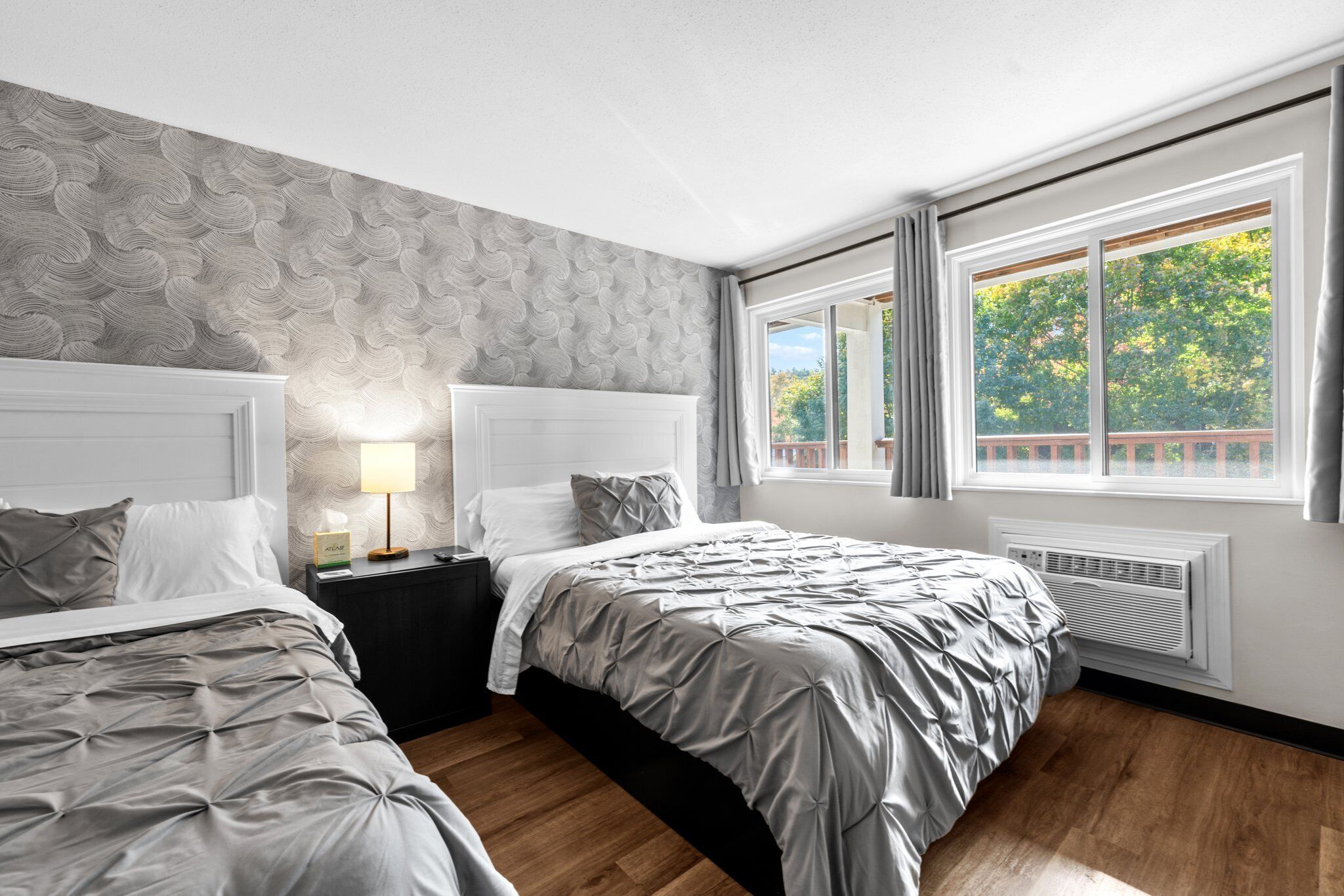 Un dormitorio moderno con dos camas, un papel tapiz estampado, una mesita de noche con lámpara y una ventana con vistas a árboles.