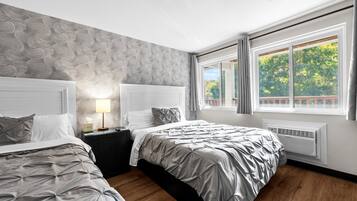 Un dormitorio moderno con dos camas, un papel tapiz estampado, una mesita de noche con lámpara y una ventana con vistas a árboles.
