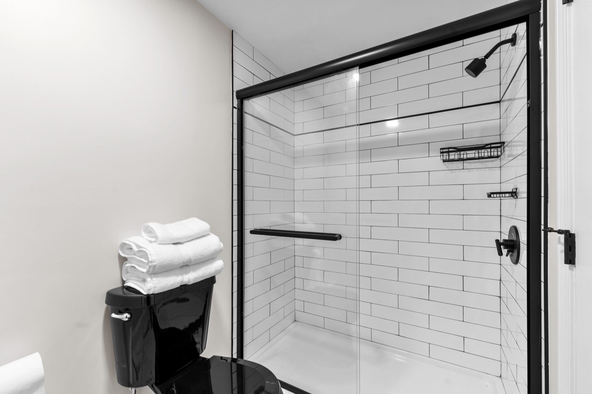Un baño moderno con ducha separada por vidrio, paredes de azulejos blancos y un inodoro negro.