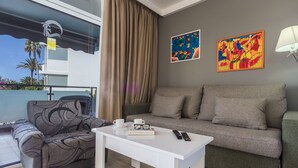 A modern living room with a grey sofa, a patterned armchair, and a white coffee table. There are two paintings on the wall, a remote control, and a pair of glasses on the table. - Alsol Patricio (San Bartolomé de Tirajana)