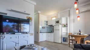 A modern kitchen with white cabinets, a microwave, and a refrigerator. There is a dining table with chairs, a TV mounted on the wall, and a small seating area with a couch. - Alsol Patricio (San Bartolomé de Tirajana)