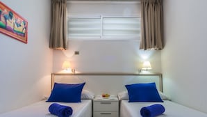 A hotel room with two beds, each with a blue pillow and a rolled towel. There is a small table with a fruit bowl and a cup between the beds. The room has a window with curtains and a painting on the wall. - Alsol Patricio (San Bartolomé de Tirajana)