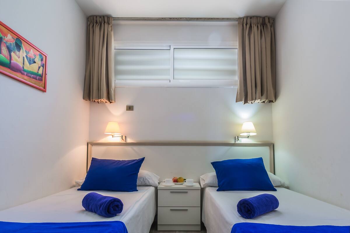 Ein Hotelzimmer mit zwei Betten, jedes mit einem blauen Kopfkissen und einem gerollten Handtuch. Zwischen den Betten steht ein kleiner Tisch mit einer Obstschale und einer Tasse. Das Zimmer verfügt über ein Fenster mit Vorhängen und ein Bild an der Wand.