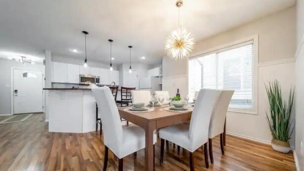 A modern kitchen with white cabinets, a dining area with a wooden table and chairs, and a large window with blinds. - Luxurious & Cozy-5bdrm Home-Minutes to WEM-WIFI (Edmonton)