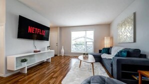 A modern living room with a flat-screen TV mounted on the wall, a white console table, a grey sofa, a round coffee table, and a large window providing natural light. - Luxurious & Cozy-5bdrm Home-Minutes to WEM-WIFI (Edmonton)