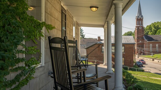 Une véranda couverte avec deux chaises en bois, un bâtiment en briques avec un clocher en arrière-plan, et une rue avec des voitures.