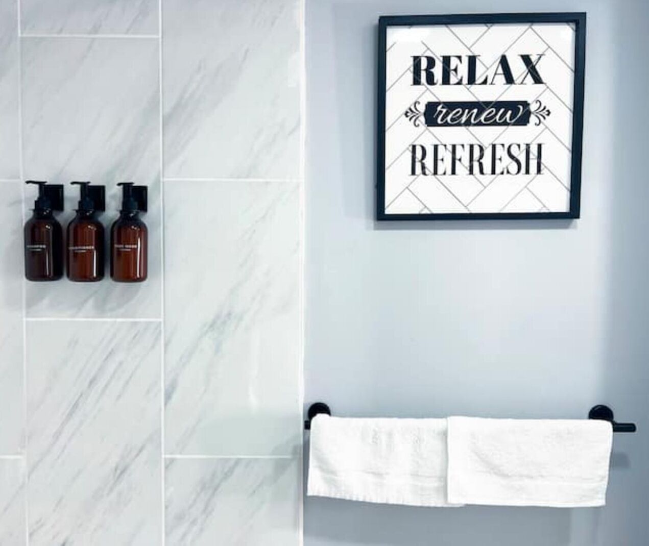 Une salle de bain avec un mur en marbre, trois bouteilles de shampoing, un panneau encadré sur lequel on peut lire « RELAXEZ-VOUS, REFAITES LE PLEIN D’ÉNERGIE, REVITALISEZ-VOUS », et un porte-serviettes avec deux serviettes.