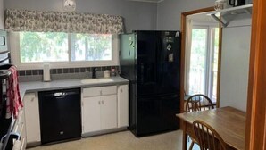 A compact kitchen with a dishwasher, a refrigerator, a window with a view of greenery, and a dining area with a table and chairs.