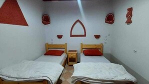 Een kamer met twee houten bedden, elk voorzien van een rood kussen en witte beddengoed. Twee nachtkastjes, elk bij een bed. De muren zijn wit, met decoratieve wanddecoraties boven de bedden.