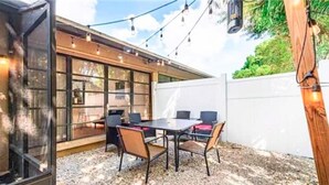 Un espace patio avec une table et des chaises, une pergola illuminée de guirlandes lumineuses et une clôture blanche.