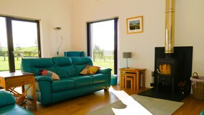 Ein Wohnzimmer mit einem türkisfarbenen Sofa, einem Holztisch, einem kleinen Beistelltisch, einem Kamin und Blick ins Freie durch große Fenster.