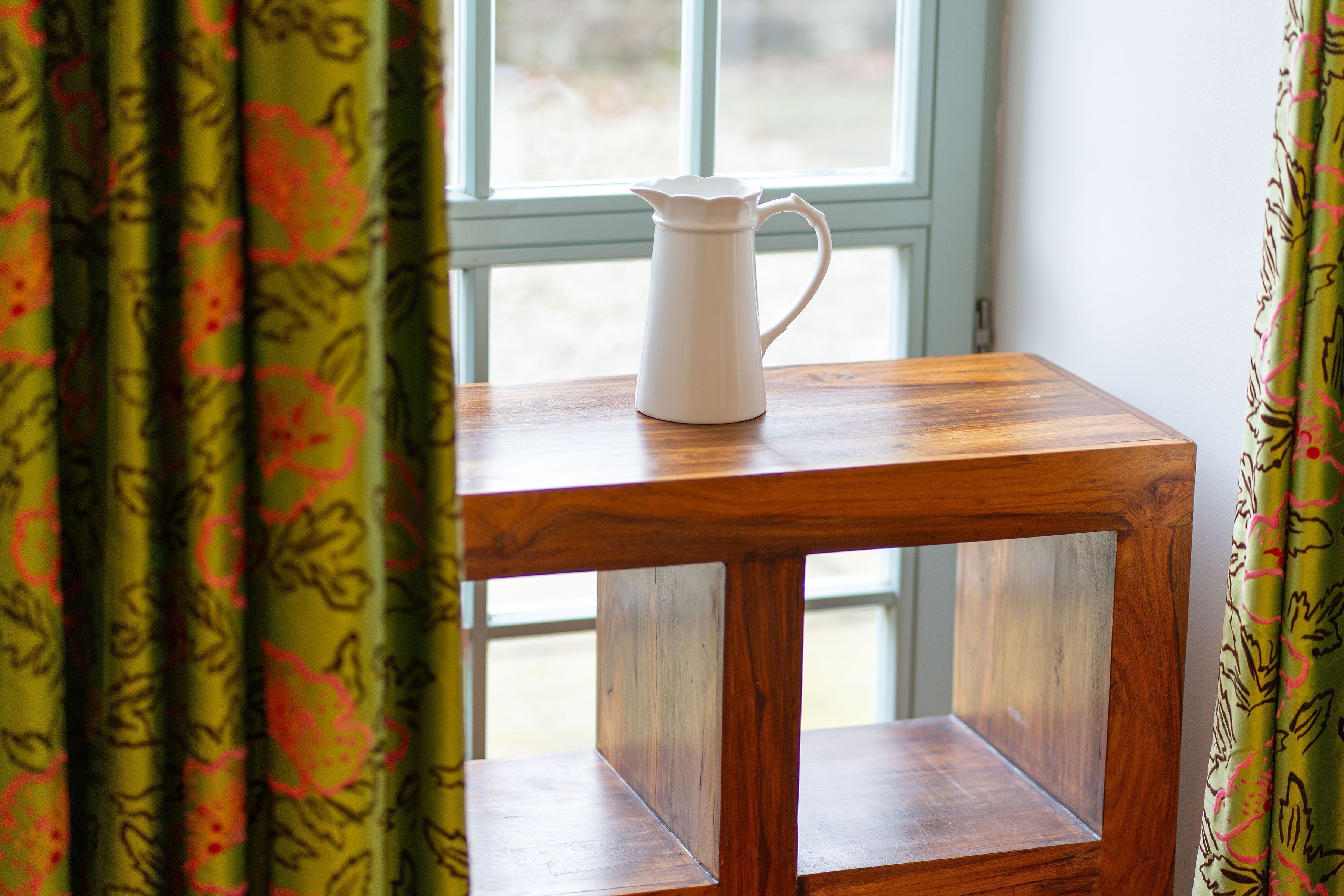 A white pitcher on a wooden table near a window with patterned curtains.