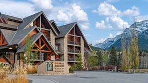 A multi-story lodge with a stone facade and wooden accents, named Grande Rockies Resort, set against a backdrop of mountains and clear skies.