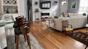 Un soggiorno moderno con un divano bianco, un tavolo da pranzo in vetro e sedie, una TV a schermo piatto montata a parete e un angolo cottura con una cantinetta per vini e mobili.