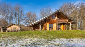 Une cabane rustique en rondins de bois, dotée d’un grand porche et d’une cheminée, entourée d’une pelouse et d’arbres.