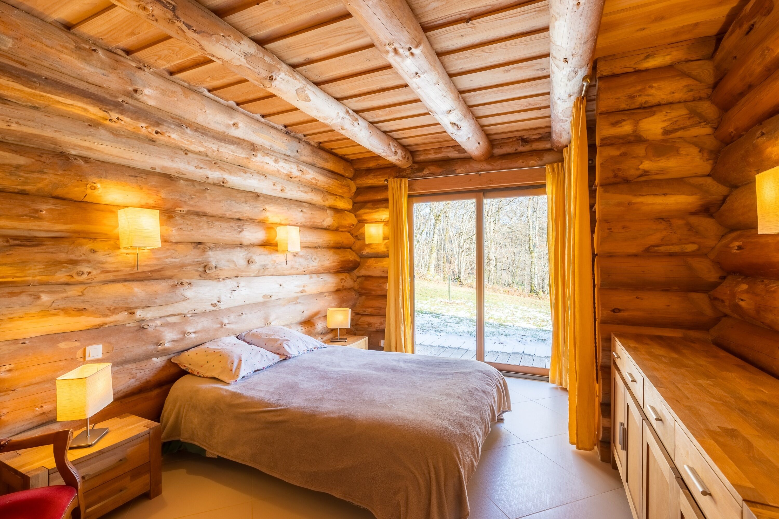 A log cabin bedroom with a bed, bedside lamps, a nightstand, and a view of the outdoors.