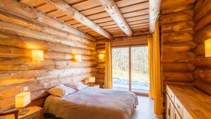 Une chambre à coucher aménagée dans un chalet en rondins, avec un lit, des lampes de chevet, une table de chevet et une vue sur l’extérieur.