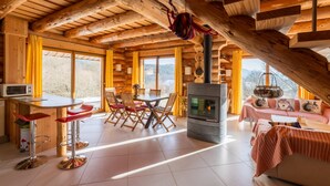 Un chalet spacieux avec un plafond haut, une cheminée centrale, un coin repas avec une table et des chaises en bois, et de grandes fenêtres offrant une vue sur l’extérieur.