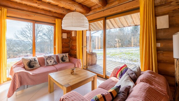 L’intérieur d’un chalet confortable, avec des murs en rondins de bois, un plafond en bois, de grandes fenêtres et un canapé agrémenté de coussins.