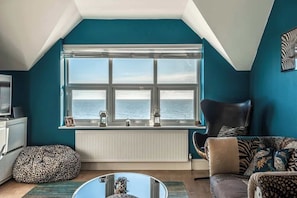 Ein Zimmer mit großem Fenster, einem Heizkörper, einem Leopardenmuster-Sitzsack, einem Ledersessel und einem gemusterten Sofa.