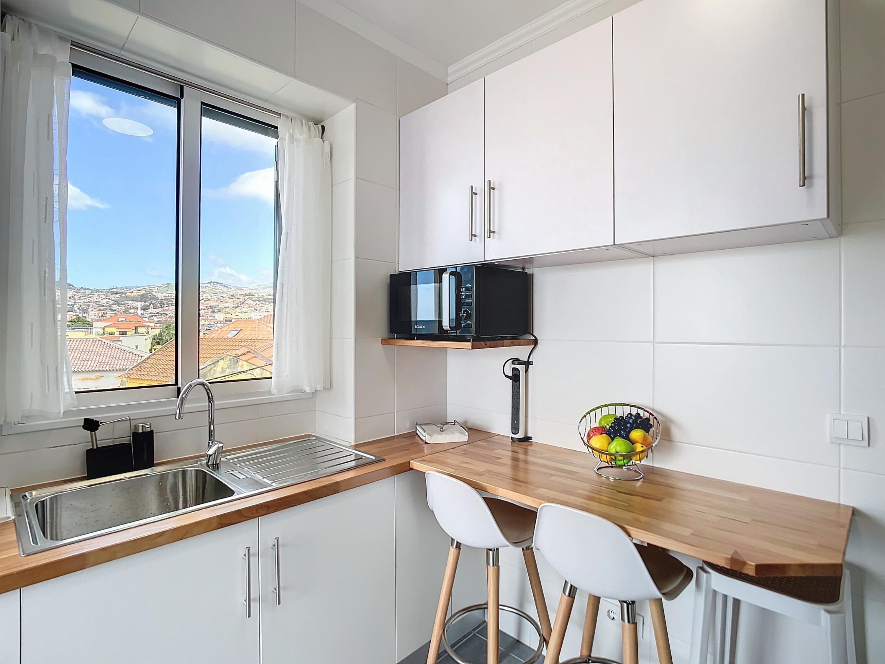 A kitchen with a window, white cabinets, a microwave, a sink, and two white chairs.