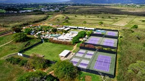 An aerial view of a sports complex with multiple tennis courts, surrounded by greenery and distant mountains.