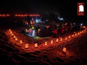 Un grupo de personas sentadas en esteras en una zona arenosa con faroles iluminados dispuestos en círculo.