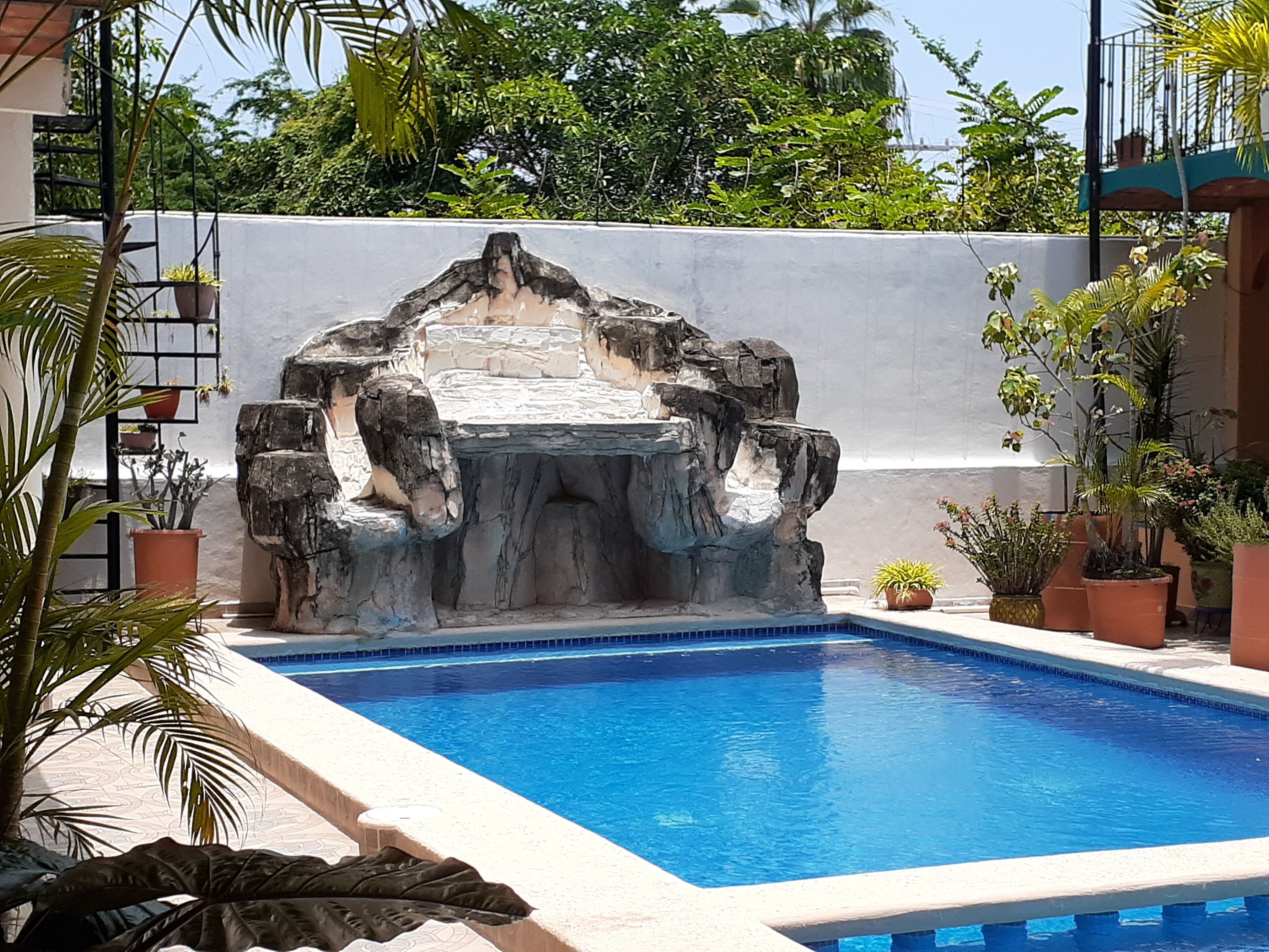 Una piscina con cascada de roca, rodeada de plantas en macetas y un muro blanco.