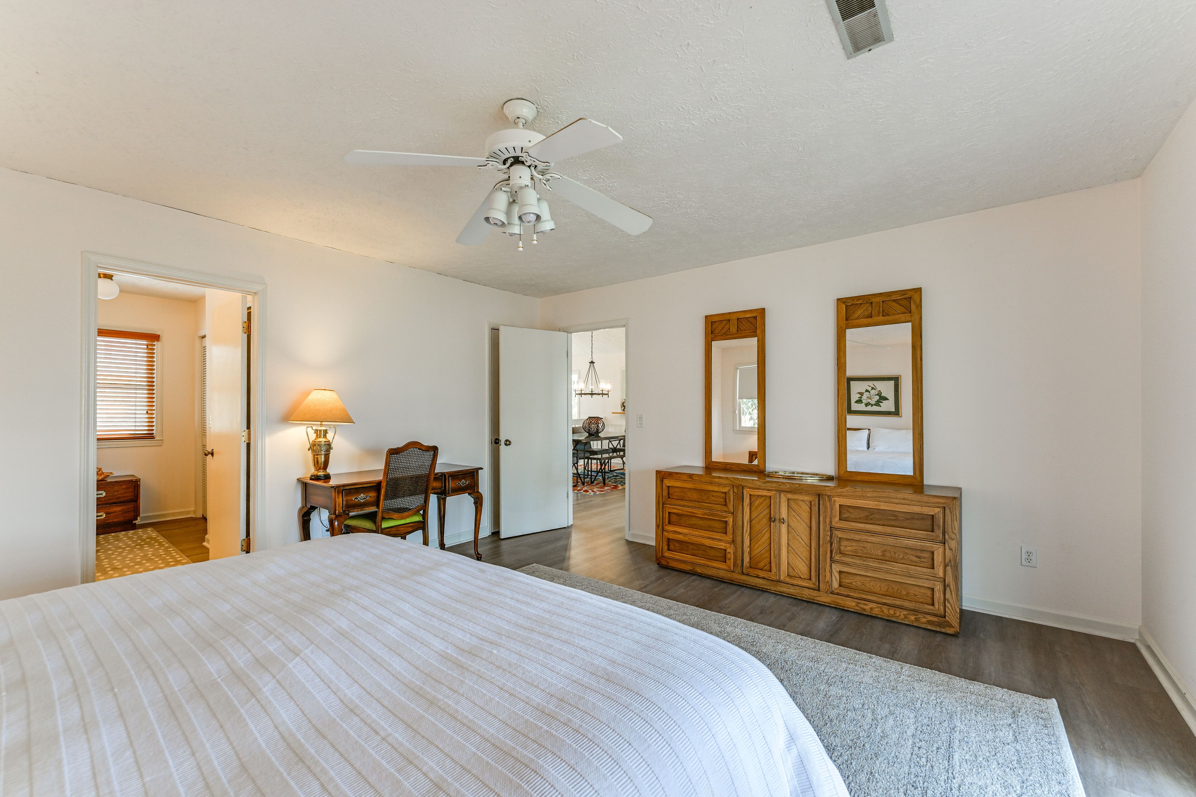 A bedroom with a large bed, a wooden dresser, a desk with a chair, and a ceiling fan.