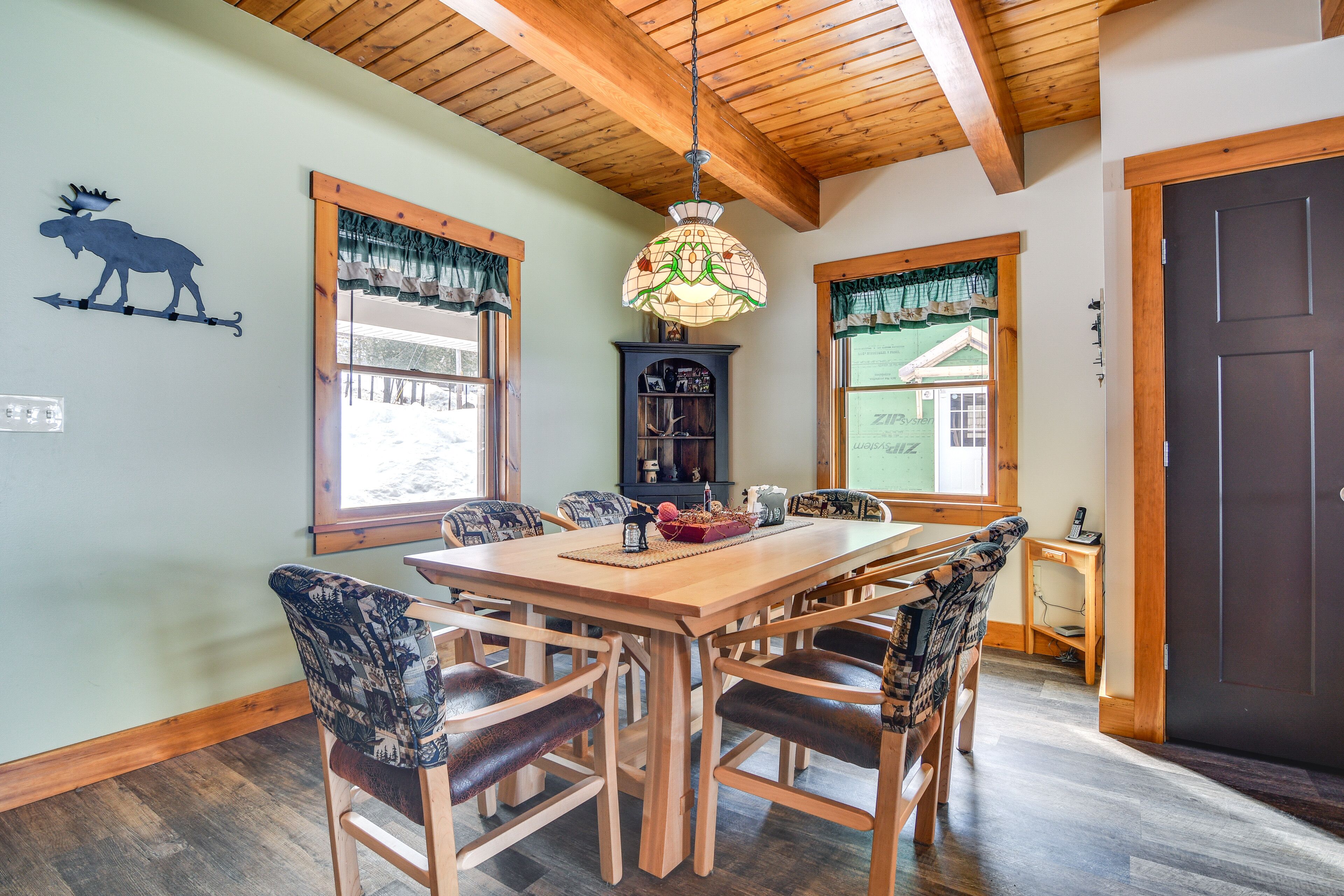 A dining area with a wooden table and chairs, a chandelier, and a view of the outdoors through windows.