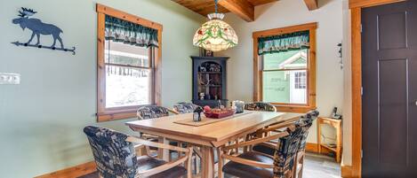 A dining area with a wooden table and chairs, a chandelier, and a view of the outdoors through windows.