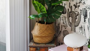 A room with a cactus-themed wallpaper, a potted monstera plant, a vintage wooden table, and a pink dresser with a white lamp. - Palme Diem! Condo Close to Downtown! (Palm Springs)