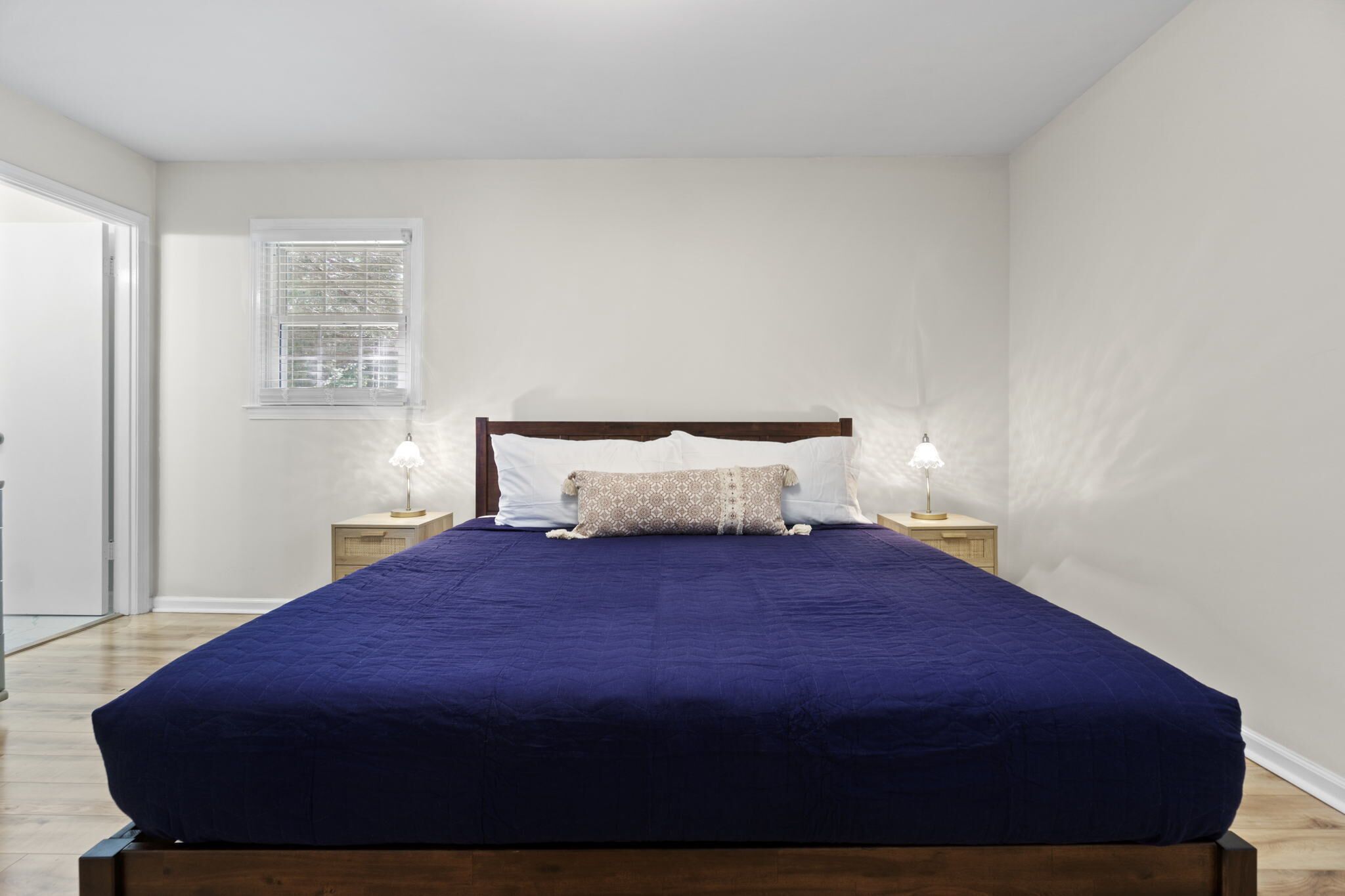 A neatly made bed with a dark blue bedspread and two pillows, flanked by two bedside tables with lamps, in a room with a window and a door.