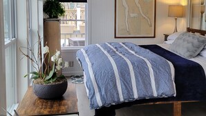 Een knus slaapkamer met een bed, een klein tafeltje met een plant, een kaart aan de muur en een raam met jaloezieën.